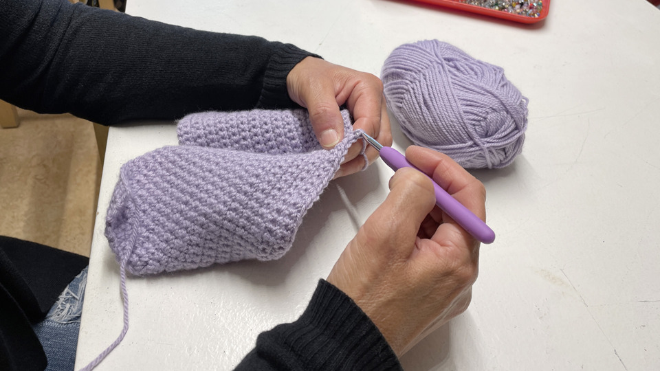 En person virkar ett lila plagg med en lila virknål. Garnet är också lila och det virkade arbetet hålls ihop.