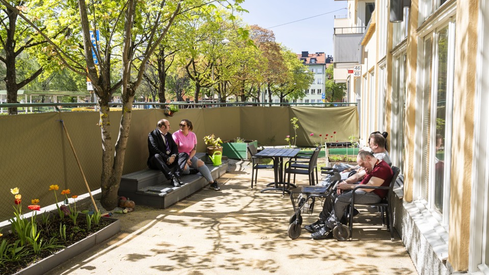 Flera personer sitter på en solig uteplats med blomplanteringar och tar det lugnt. Uteplatsen är omgiven av träd.
