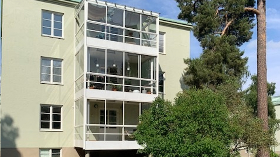 Husfasad med tre våningar och en inglasad balkong på varje våning. En stor tall utanför huset.