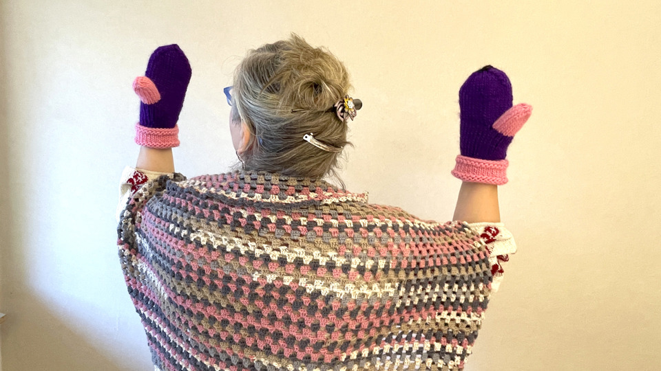 En person visar upp ett par stickade handskar i lila med rosa detaljer på tummen och muddarna. Personen bär också en virkad sjal i flera färger.