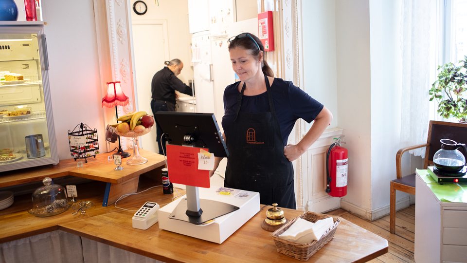 En kvinna står framför kassan i ett kafé. i bakgrunden står en man och arbetar vid en bänk.