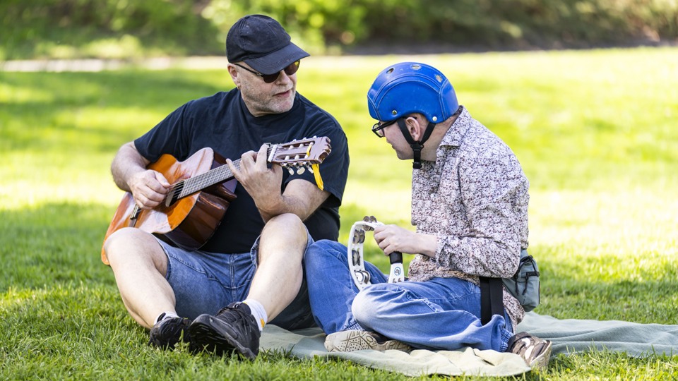 Två män sitter i gräset. Den ena mannen spelar gitarr, den andre mannen spelar tamburin.