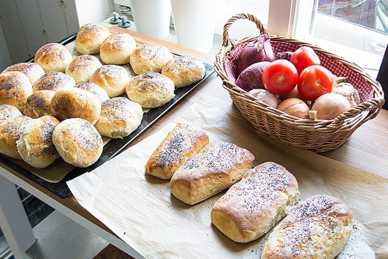 Nybakade småbröd på en plåt och vid sidan om på ett bakplåtspapper samt korg med lök och tomater.