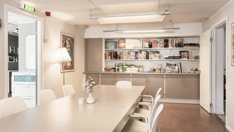 Ljust rum med vita väggar, skåp och hyllpr och avlångt vitt bord och vita stolar.