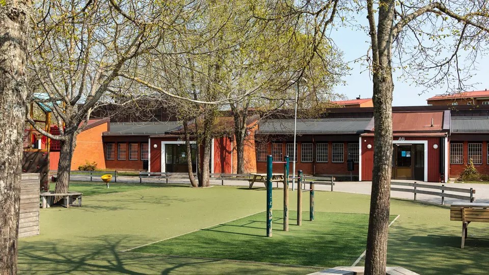 En röd skolbyggnad i en våning med en gård med klätterställning och snurrstol.