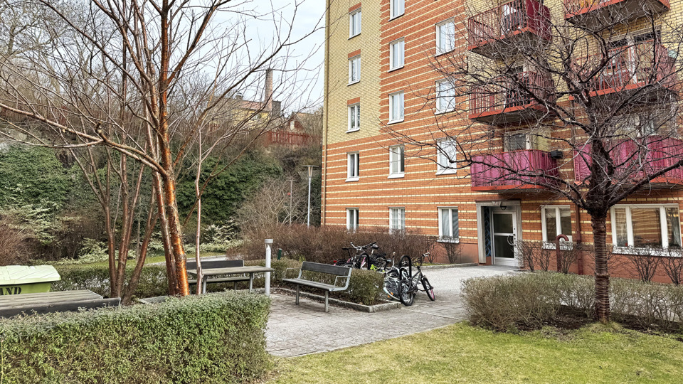 Orange och gul tegelbyggnad med röda balkonger. Bänkar och träd utanför.