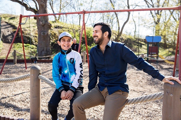 En man och en pojke sitter och skrattar i en park och solen skiner.