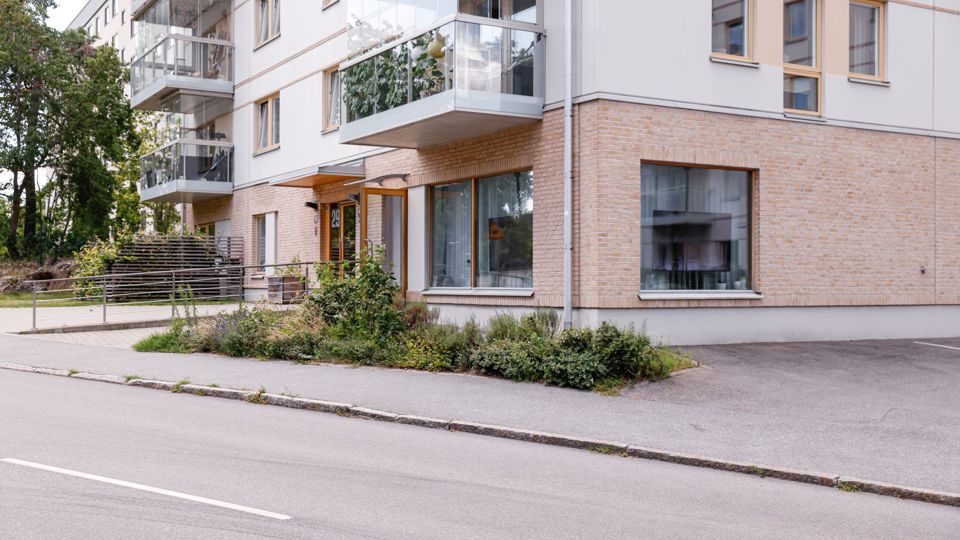 Ljust flervåningshus omgivet av buskar intill en väg, foto.