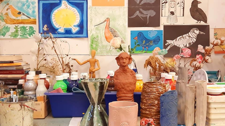 Konstnärlig arbetsyta med keramikföremål, färgburkar och skulpturer på ett bord. Bakom syns en vägg täckt av målningar med fåglar och naturmotiv.