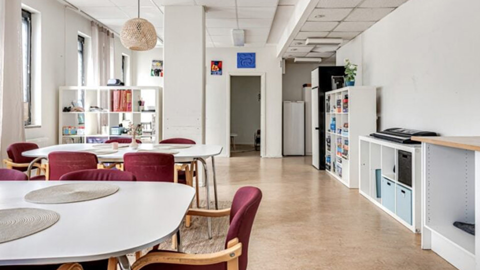 Ljust avlångt allrum med vita väggar. Det finns bord, stolar och hyllor. 