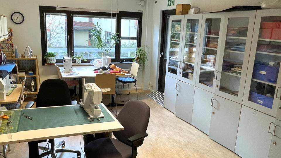 En syverkstad med arbetsbord och förvaringsskåp. I bakgrunden syns växter utanför fönstren och en symaskin på ett av borden.