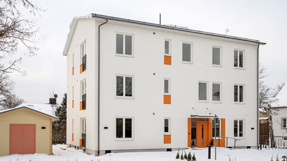 Vit byggnad i tre våningar med många fönster och franska balkonger på gaveln. Liten trädgård och ett förråd intill huset.
